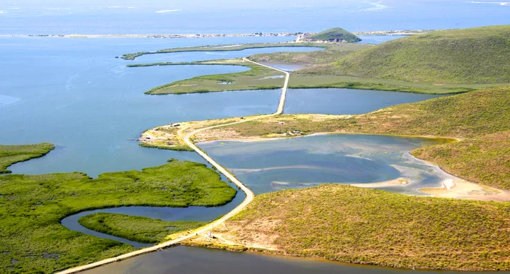 Vista aérea de una carretera elevada que atraviesa esteros y lagunas costeras hacia una barra de arena frente al mar abierto