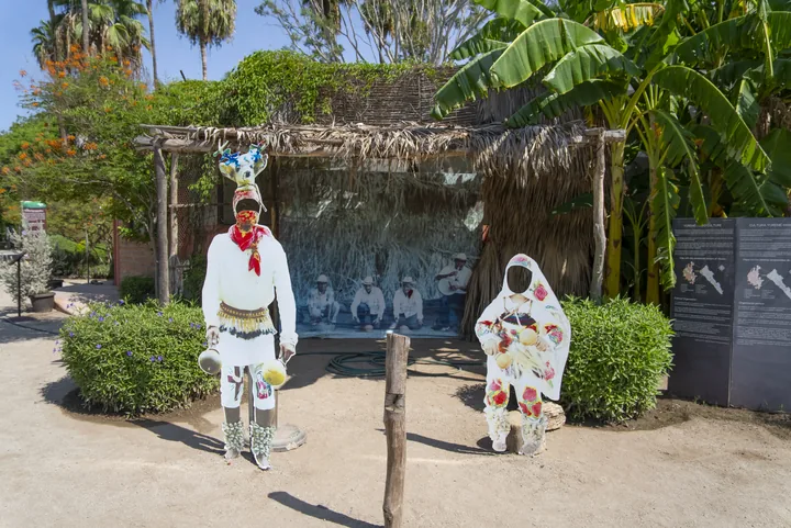 Figuras a escala real de un danzante de venado yoreme y una mujer con manto bordado, frente a una casa de palma y paneles informativos sobre la cultura yoreme-mayo