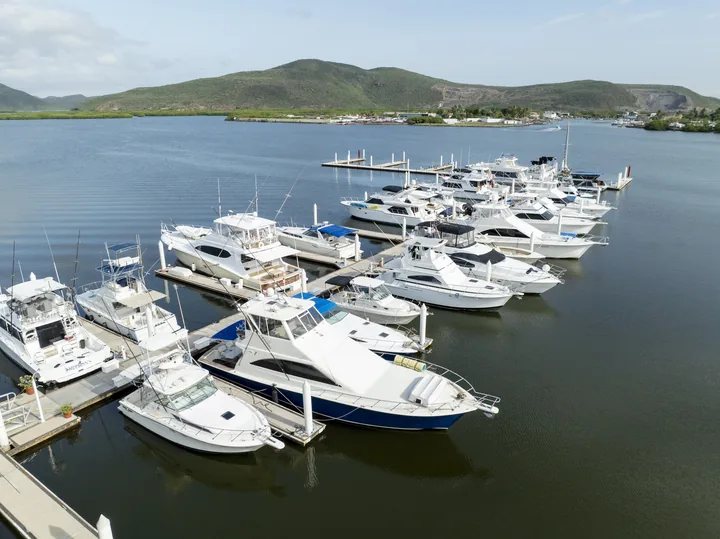 Vista aérea de una marina con embarcaciones deportivas atracadas, con cerros verdes y manglar al fondo
